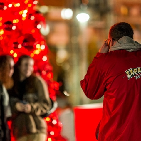Άνδρας ντυμένος στα κόκκινο βγάζει φωτογραφία δύο κοπέλες μπροστά από ένα κόκκινο χριστουγεννιάτικο δέντρο
