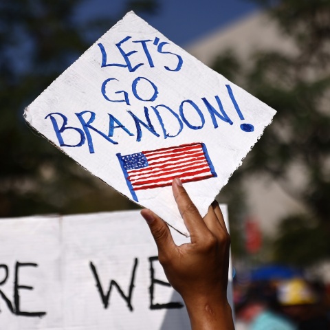 Χέρι που κρατά πινακίδα στην οποία αναγράφεται η φράση Let's Go Brandon