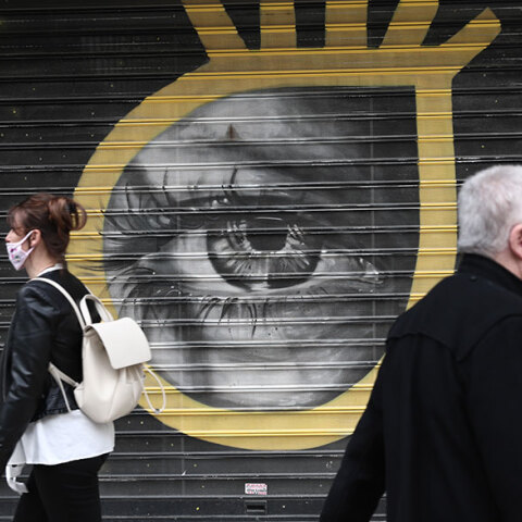 Πολίτες με μάσκα για κορωνοϊό μπροστά από γκράφιτι για την πανδημία Covid-19