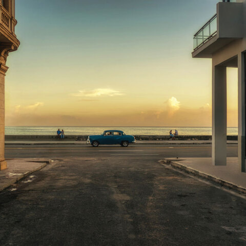Ένα μπλε αυτοκίνητο φαίνεται στο βάθος της φωτογραφίας, ανάμεσα σε δύο κτήρια και με τη θάλασσα στο φοντο, ώρα ηλιοβασιλέματος