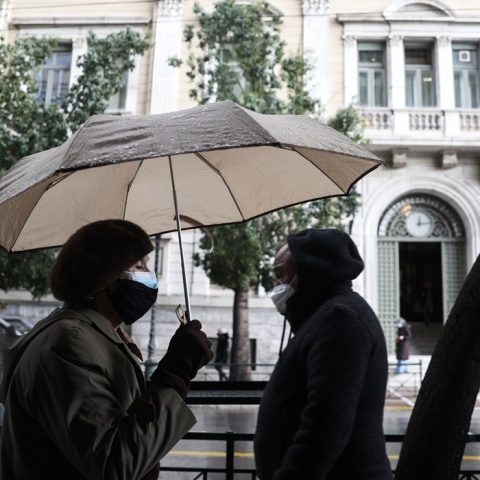 Γυναίκα περπατά κρατώντας την ομπρέλα της - Στο βάθος άνδρας έχει φορέσει την κουκούλα για να προστατευτεί από τη βροχή