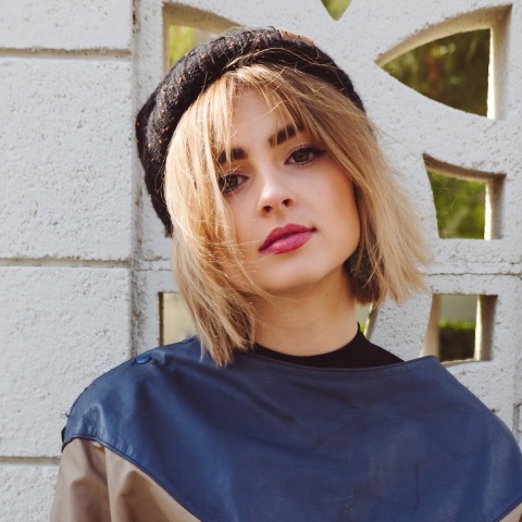 Ξανθιά κοπέλα με φούτερ και μπερέ ποζάρει μπροστά από λευκό τοίχο με σκαλιστό παραθυράκι