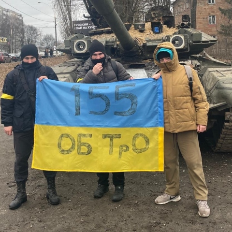 Τρεις νεαροί άνδρες κρατούν σημαία της Ουκρανίας μπροστά από τανκ