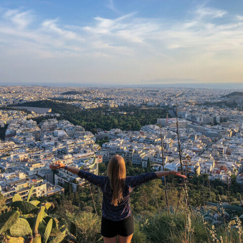 Κοπέλα στέκεται όρθια με γυρισμένη πλάτη με φόντο την Αθήνα από ψηλά