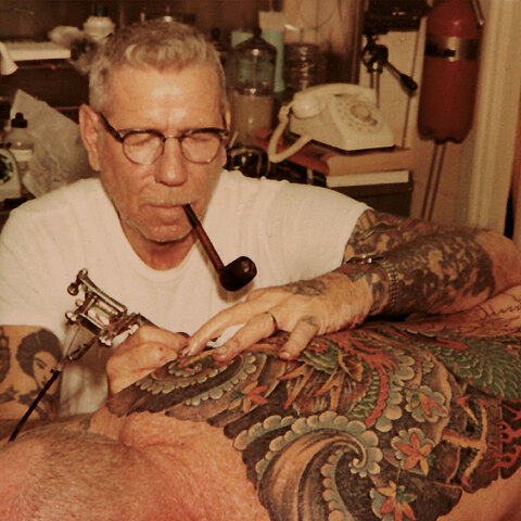 Norman Collins tattooing back with pipe in mouth
