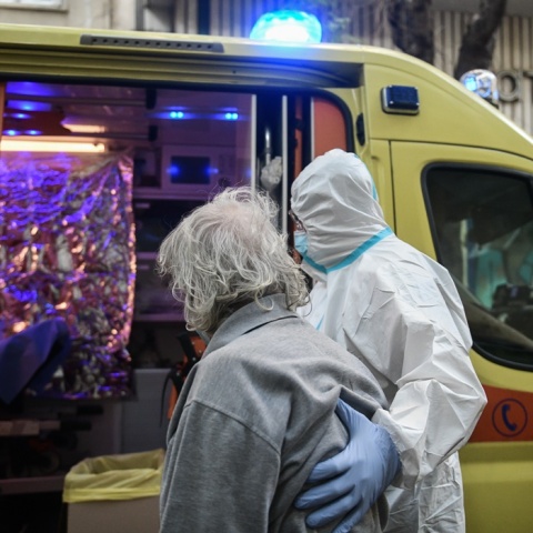 Ηλικιωμένη με κορωνοϊό και ζακέτα ριγμένη στους ώμους μεταφέρεται από άνδρα με ειδική στολή στο νοσοκομείο