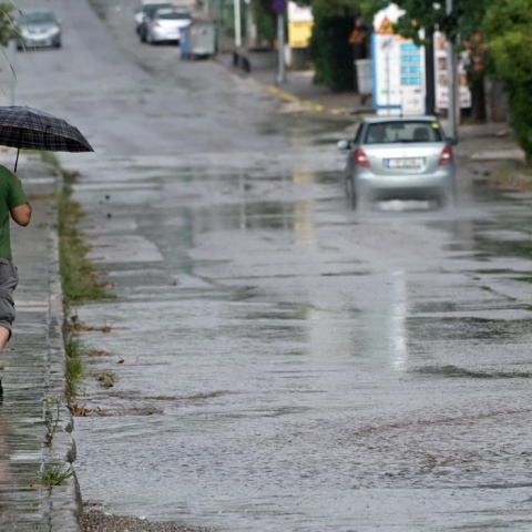 Πολίτης με ομπρέλα σε πεζοδρόμιο και δίπλα του αυτοκίνητο διασχίζει πλημμυρισμένο δρόμο