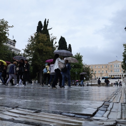 Πολίτες στο κέντρο της Αθήνας κρατούν ομπρέλες διασχίζοντας τον δρόμο που οδηγεί στην πλατεία Συντάγματος