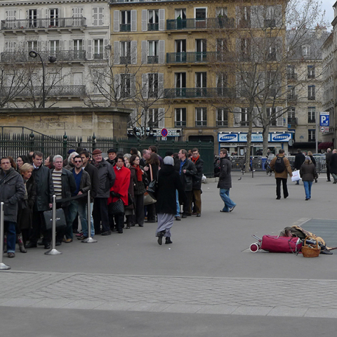 Άνθρωποι που περιμένουν σε μια ουρά και μια άστεγη γυναίκα κάθεται κοντά τους στην πλατεία Madeleine στο Παρίσι