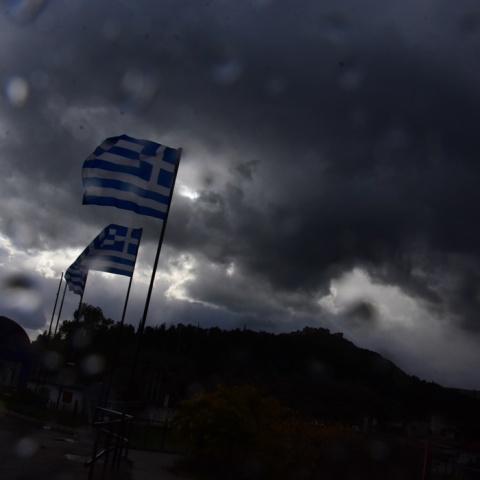 Σταγόνες βροχής πάνω σε τζάμι και στο φόντο ελληνικές σημαίες 