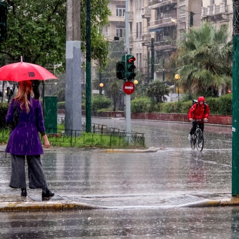 Γυναίκα με ομπρέλα περιμένει σε διάβαση πεζών την ώρα που ποδηλάτης με αδιάβροχο κινείται στους δρόμους της Αθήνας