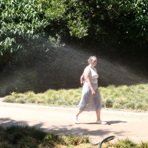 Γυναίκα περπατά σε συνθήκες καύσωνα σε περιβάλλον όπου ποτιστικό δροσίζει φυτά πετώντας νερό