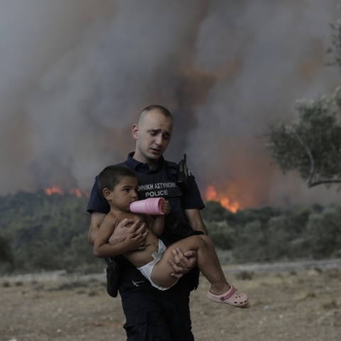 Αστυνομικός μεταφέρει στην αγκαλιά του παιδί ενώ πίσω τους μαίνεται η φωτιά στα Δερβενοχώρια