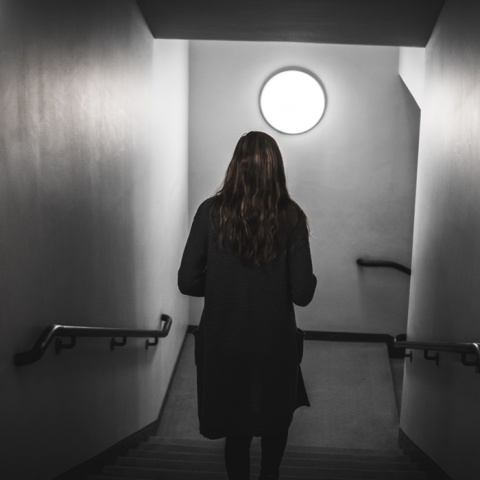 Γυναίκα περπατά με την πλάτη γυρισμένη στον φακό προς πόρτα που βρίσκεται σε κατηφορικό σκοτεινό διάδρομο