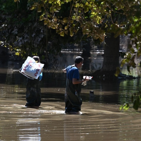 Άνδρες με προστατευτικά περπατούν σε πλημμυρισμένο δρόμο και κουβαλούν πράγματα