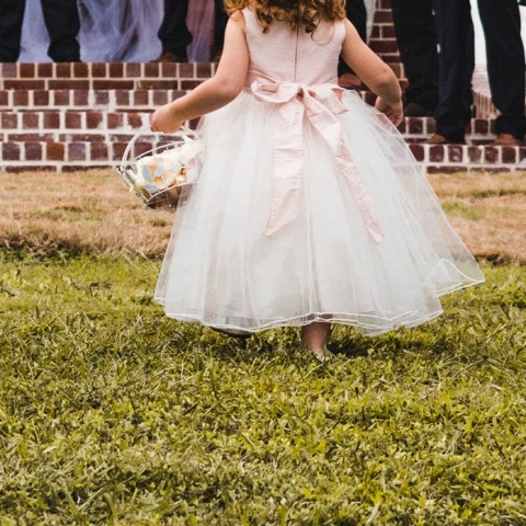 Κοριτσάκι με φόρεμα που παραπέμπει σε γάμο διασχίζει σημείο με γρασίδι και κρατά στα χέρια του καλαθάκι