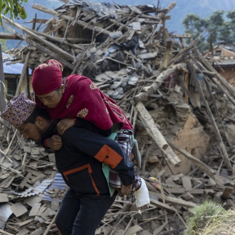 Άνδρας μεταφέρει γυναίκα στην πλάτη του ενώ πίσω τους υπάρχουν χαλάσματα από τον σεισμό στο Νεπάλ