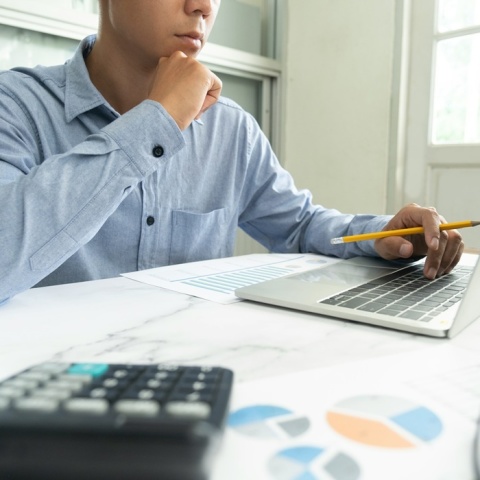 Άτομο μπροστά από λάπτοπ με χαρτιά που απεικονίζουν γραφήματα κάνει υπολογισμούς