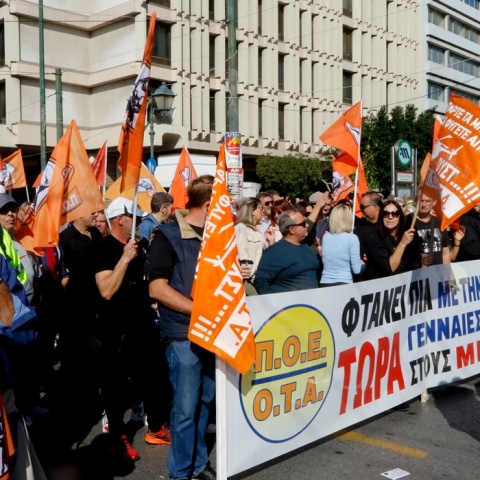 Άτομα που συμμετέχουν σε συγκέντρωση στο κέντρο της Αθήνας κρατώντας σημαίες και πανό της ΠΟΕ-ΟΤΑ