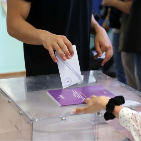 Χέρι που ρίχνει φάκελο στην κάλπη και άλλο χέρι που βγάζει το βιβλίο από τη σχισμή της κάλπης