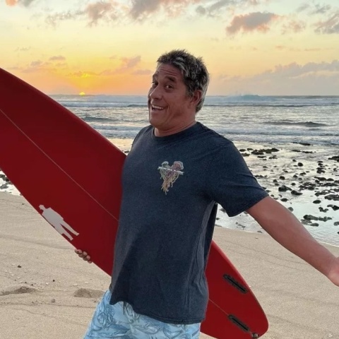 Ο Ταμάγιο Πέρι ποζάρει σε παραλία με τη σανίδα του σερφ ανά χείρας