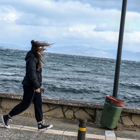 Γυναίκα που φορά μπουφάν περπατά εν μέσω δυνατού αέρα σε παραλιακό μέρος