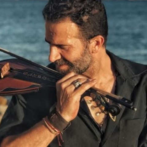 Ο Ηλίας Παλιουδάκης μπροστά από τη θάλασσα κρατώντας παραδοσιακό μουσικό όργανο