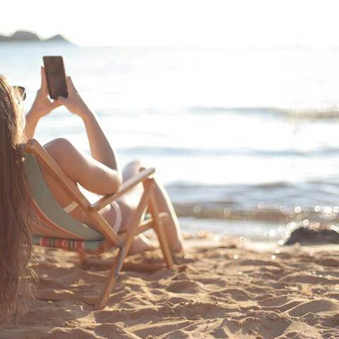 Γυναίκα σε ξύλινη καρέκλα στην παραλία κοιτά το κινητό της τηλέφωνο