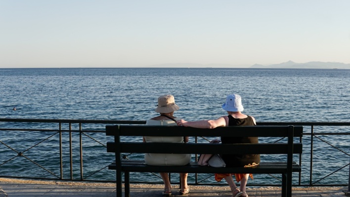 Ηλικιωμένες κυρίες με πλάτη στο φακό φορούν το καπέλο τους και κάθονται σε παγκάκι με θέα τη θάλασσα