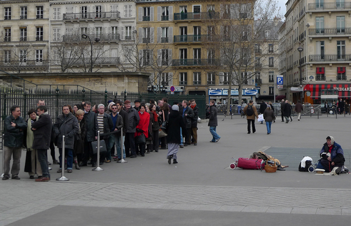 Άνθρωποι που περιμένουν σε μια ουρά και μια άστεγη γυναίκα κάθεται κοντά τους στην πλατεία Madeleine στο Παρίσι