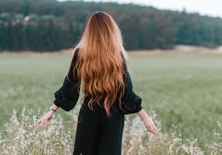 Κοπέλα με μακριά μαλλιά και γυρισμένη πλάτη περπατάει σε χωράφι αγγίζοντας τα φυτά