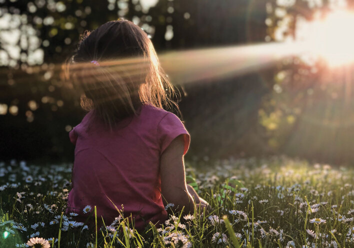 Κοριτσάκι γυρισμένο πλάτη κάθεται στο γρασίδι με φόντο δέντρα και ήλιο