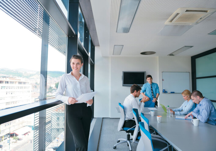 Κοπέλα όρθια σε χώρο γραφείου, στο βάθος άντρες καθιστοί γύρω από τραπέζι