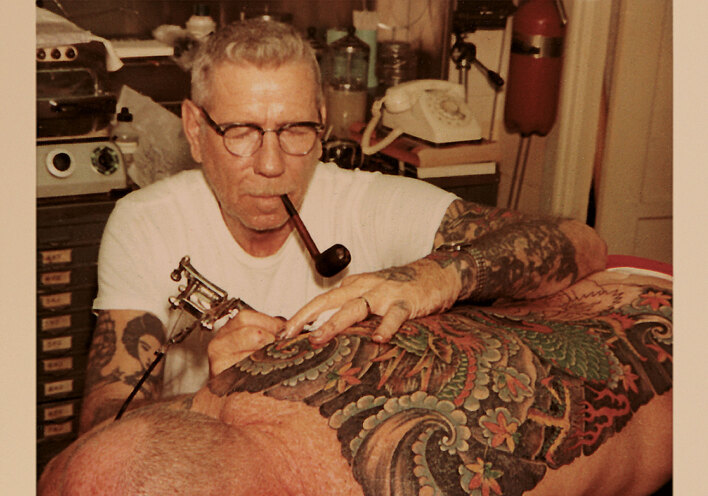 Norman Collins tattooing back with pipe in mouth