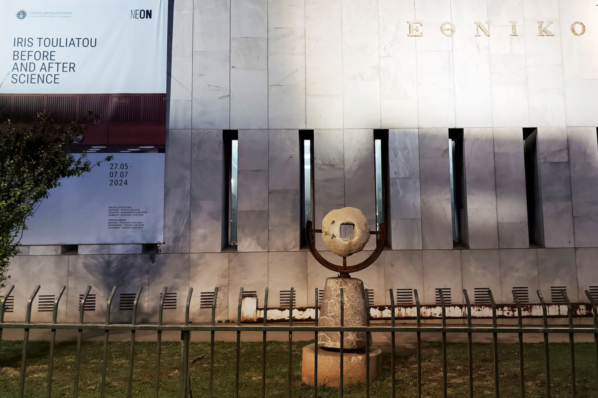 «Progress in Optics», 2024. Ένας προβολέας που προστέθηκε στην υπάρχουσα υποδομή φωτισμού της σήμανσης της πρόσοψης του κτιρίου, σκιαγραφεί το ελλείπον Ν που αφαιρέθηκε σύμφωνα με τις ορθογραφικές μεταρρυθμίσεις της ελληνικής γλώσσας