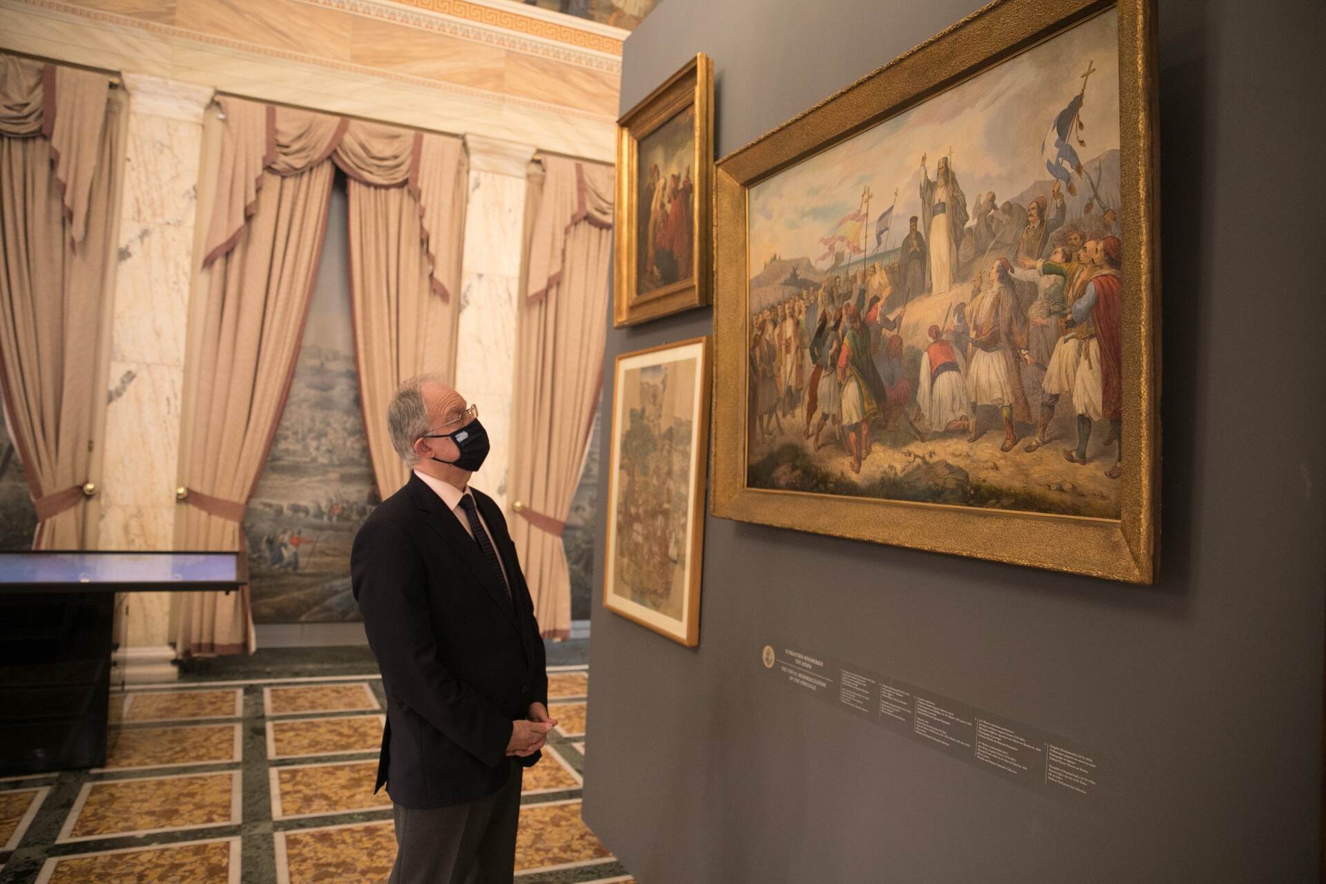 Ο Πρόεδρος της Βουλής Κωνσταντίνος Τασούλας μπροστά σε έργα απεικόνισης της Επανάστασης. Στο βάθος φαίνεται ένα από τα παράθυρα με τα ριντό που δείχνουν πώς φαινόταν η σημερινή πλατεία Συντάγματος την εποχή που κτίστηκε η Βουλή