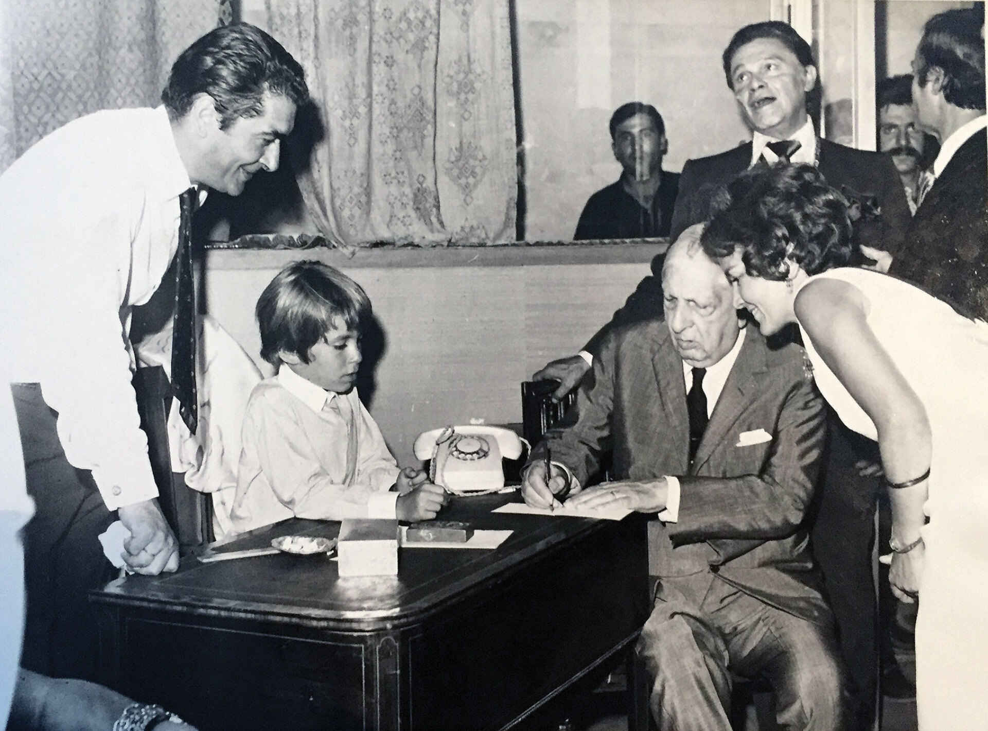 Εγκαίνια έκθεσης του De Chirico, δεκαετία του ’70. Διακρίνονται αριστερά ο Τάσος Ζουμπουλάκης με το γιο του Θοδωρή, εν αναμονή αυτογράφου από τον καλλιτέχνη κάτω από το βλέμμα της Πέγκυς και του Ιόλα (στο βάθος). Ήρθε στα εγκαίνια, τον φιλοξενήσαν μια εβδομάδα μαζί με τη γυναίκα του στην Ανάβυσσο.