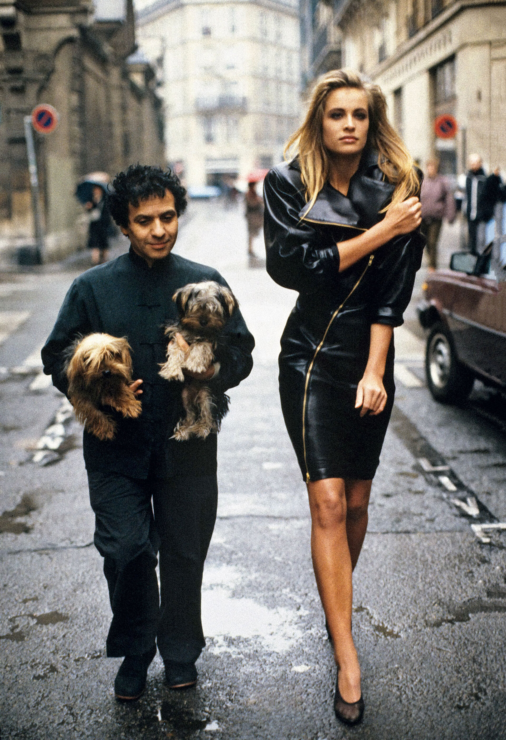Ο σχεδιαστής Azzedine Alaïa περπατά στους δρόμους του Παρισιού έχοντας αγκαλιά τα δυο του γιόρκσαϊρ τερριέ, Πατατούφ και Γουάμπο. Στο πλάι του η ολλανδέζα καλλονή Φρεντερίκ βαν ντερ Ουάλ, φορώντας το εμβληματικό του μαύρο δερμάτινο φόρεμα με το φερμουάρ, 1986 © Arthur Elgort / Conde Nast via Getty Images