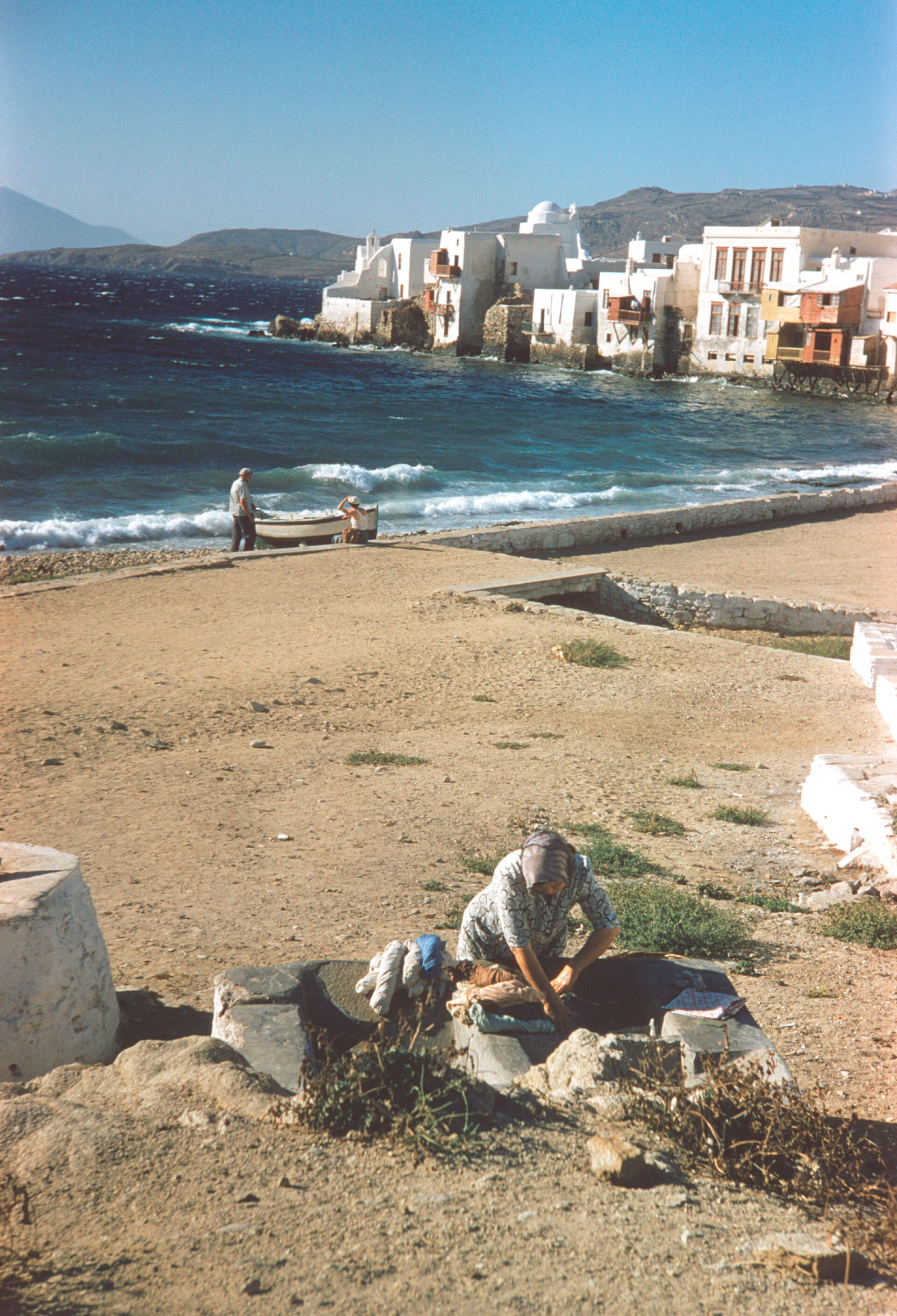 Μύκονος 1957. Μια γυναίκα βάζει μπουγάδα σε πηγάδι. Πίσω η Μικρή Βενετία
