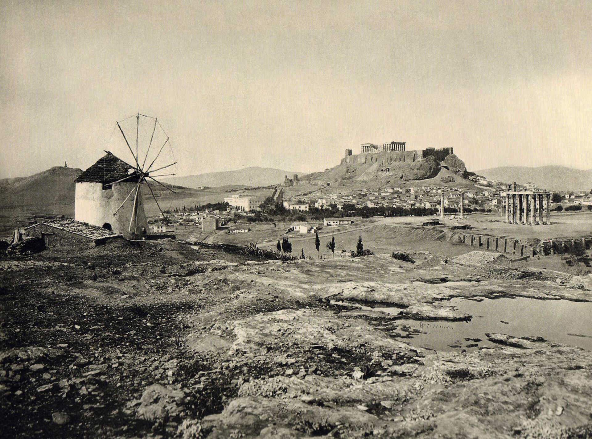 Όψη της Ακρόπολης και των Στύλων του ναού του Ολυμπίου Διός από το λόφο του Μετς. Αριστερά διακρίνεται ο μύλος Αργυρίου, ο τελευταίος ανεμόμυλος που είχε απομείνει στην περιοχή, 1869 | Φωτογραφία του Αμερικανού δημοσιογράφου Γουίλιαμ Τζέιμς Στίλμαν, 1828-1901