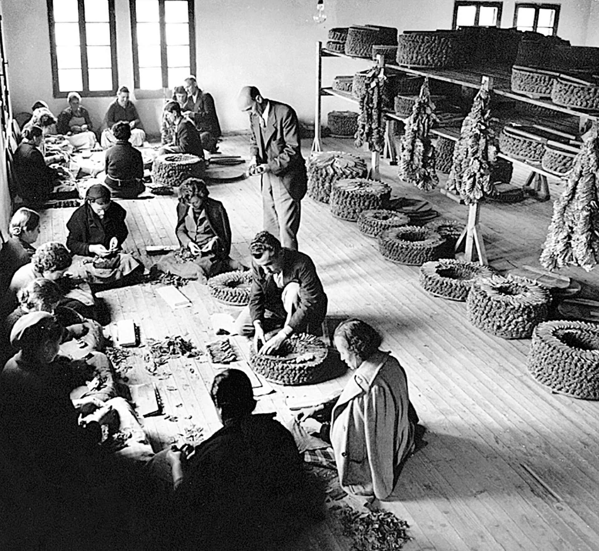 Ο ντεγκτσής, ο εξειδικευμένος τεχνίτης-δεματοποιός επί το έργον. Γύρω του οι πασταλτζούδες –  οι καπνεργάτριες βοηθοί του, που ομαδοποιούν τα καπνόφυλλα κατά σχήμα και μέγεθος πριν του τα παραδώσουν για να τα προσθέσει στο δέμα. Μουσείο Καπνού Δήμου Καβάλας. Ιδιωτική Συλλογή Βασίλη Θασίτη ΑΒΕ 115/2005/1934.