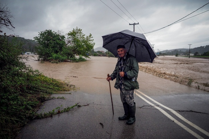 Ηλικιωμένος άνδρας με μαγκούρα και ομπρέλα μπροστά από δρόμο που έχει πλημμυρίσει στην Εύβοια