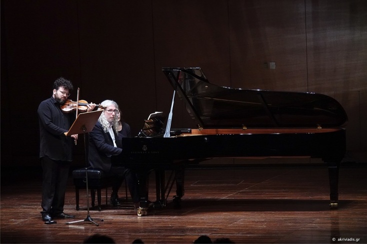 Ένας βιολιστής στα μαύρα παίζει βιολί όρθιος. Δύο άτομα κάθονται δίπλα του σε μαύρο πίανο με ούρα, σε ζωντανή παράσταση