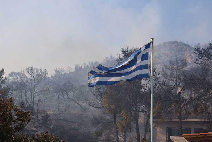Ελληνική σημαία στον ιστό της με φόντο τα καμένα της Σαλαμίνας