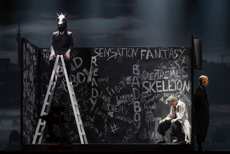 Σκηνικό με σκάλα και άντρα που κάθεται στην κορυφή της με κεφάλι αλόγου, γυναίκα καθισμένη σε σκαμπό και άνδρα όρθιο δίπλα της