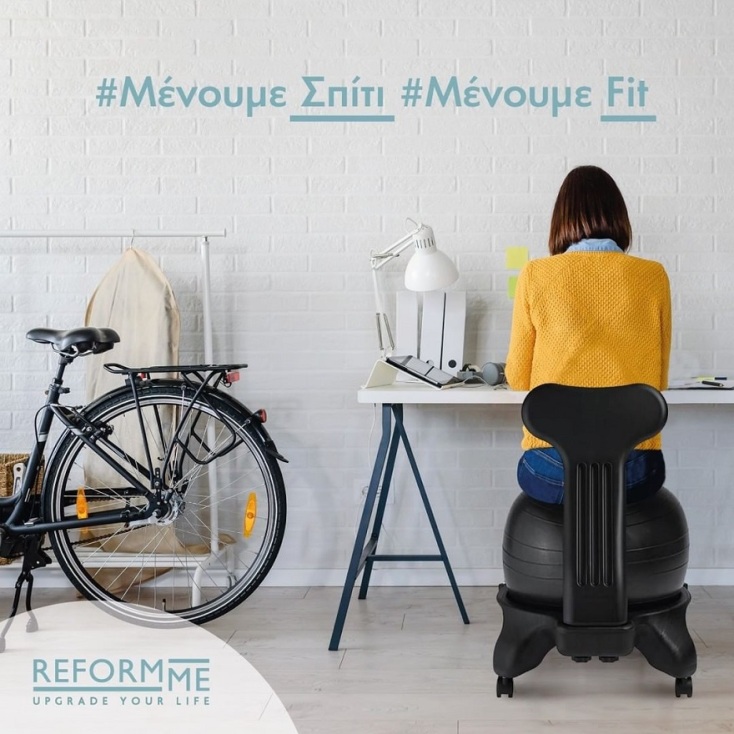Γυναίκα καθισμένη σε καρέκλα-μπάλα Fit-Chair® μπροστά σε γραφείο με γυρισμένη την πλάτη και ποδήλατο δίπλα της