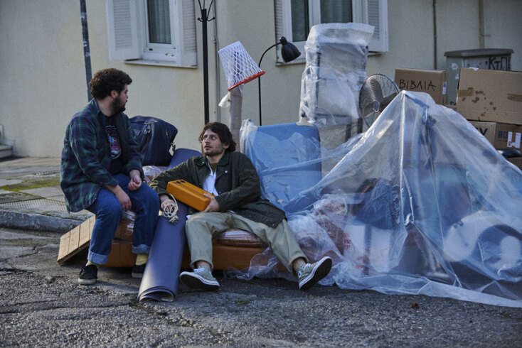 Δύο άντρες καθισμένοι ανάμεσα σε κούτες και έπιπλα στην άκρη του δρόμου