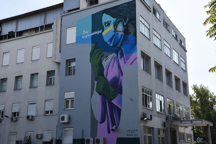 Στο γκράφιτι για τον κορωνοϊό στο Γενικό Κρατικό Νοσοκομείο Νίκαιας απεικονίζεται μια εργαζόμενη στον χώρο της Υγείας να φορά προστατευτική μάσκα και γάντια για την Covid-19