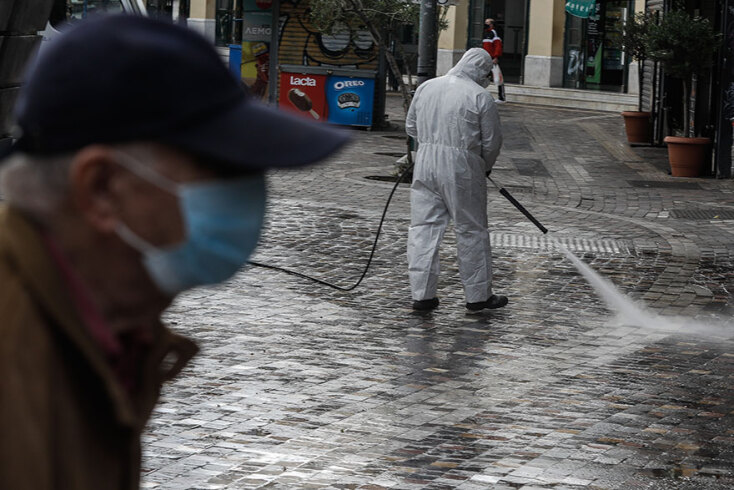 Πολίτης με μάσκα για τον κορωνοϊό περνά πίσω από υπάλληλο του Δήμου Αθηναίων που απολυμαίνει στο Μοναστηράκι, την πρώτη ημέρα του νέου lockdown
