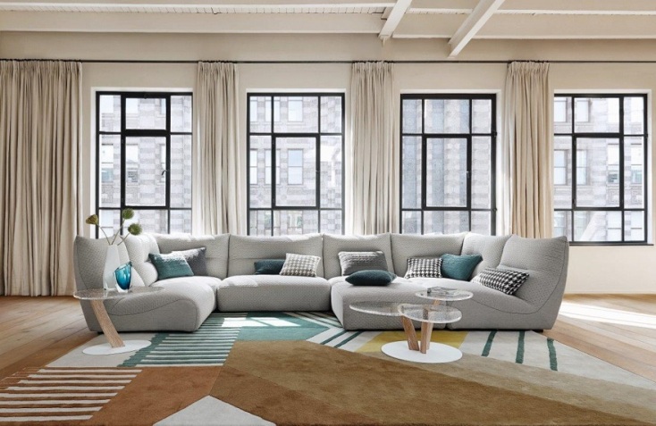 Φωτεινό σαλόνι με μεγάλα παράθυρα, λευκές κουρτίνες, μεγάλο γκρι γωνιακό καναπέ με καρό μαξιλάρια και μπεζ χαλί